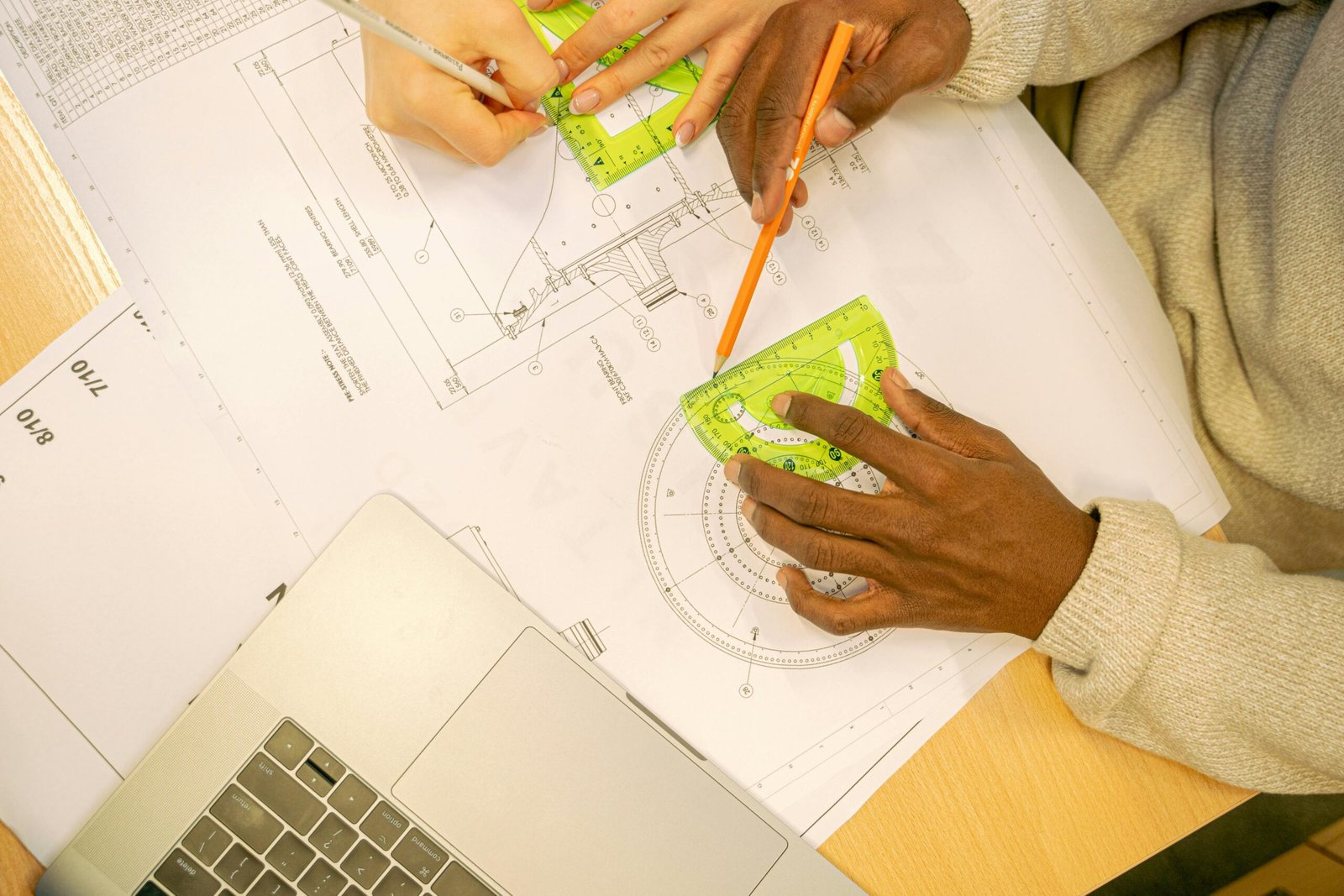Hands collaborating on a blueprint drawing with a protractor and laptop, showcasing teamwork and design precision.